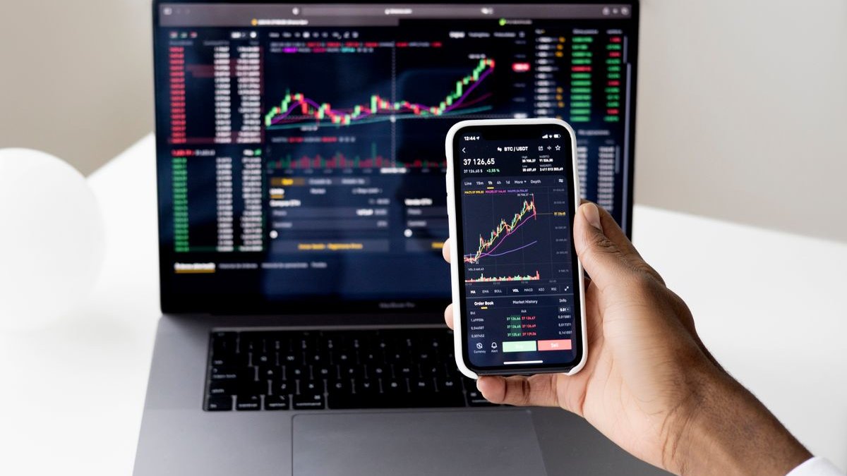 Trade Republic Konditionen: Hand hält Smartphone mit Kurschart vor Laptop